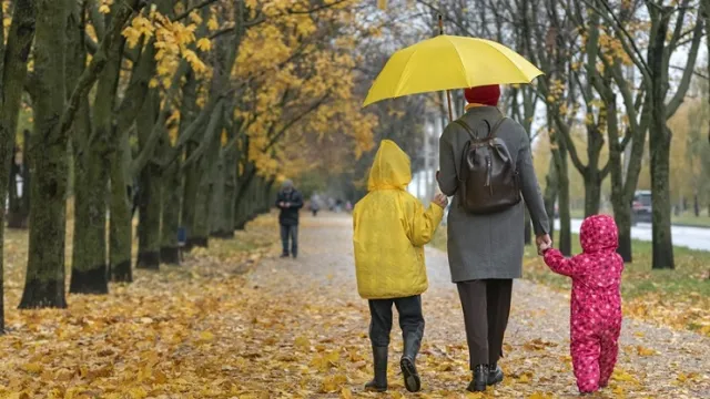 Mom with son and daughter walking along an alley with umbrella. Falling leaves. Family walk with children. Back view Mom with son and daughter walking along an alley with umbrella. Falling leaves. Family walk with children. Back view